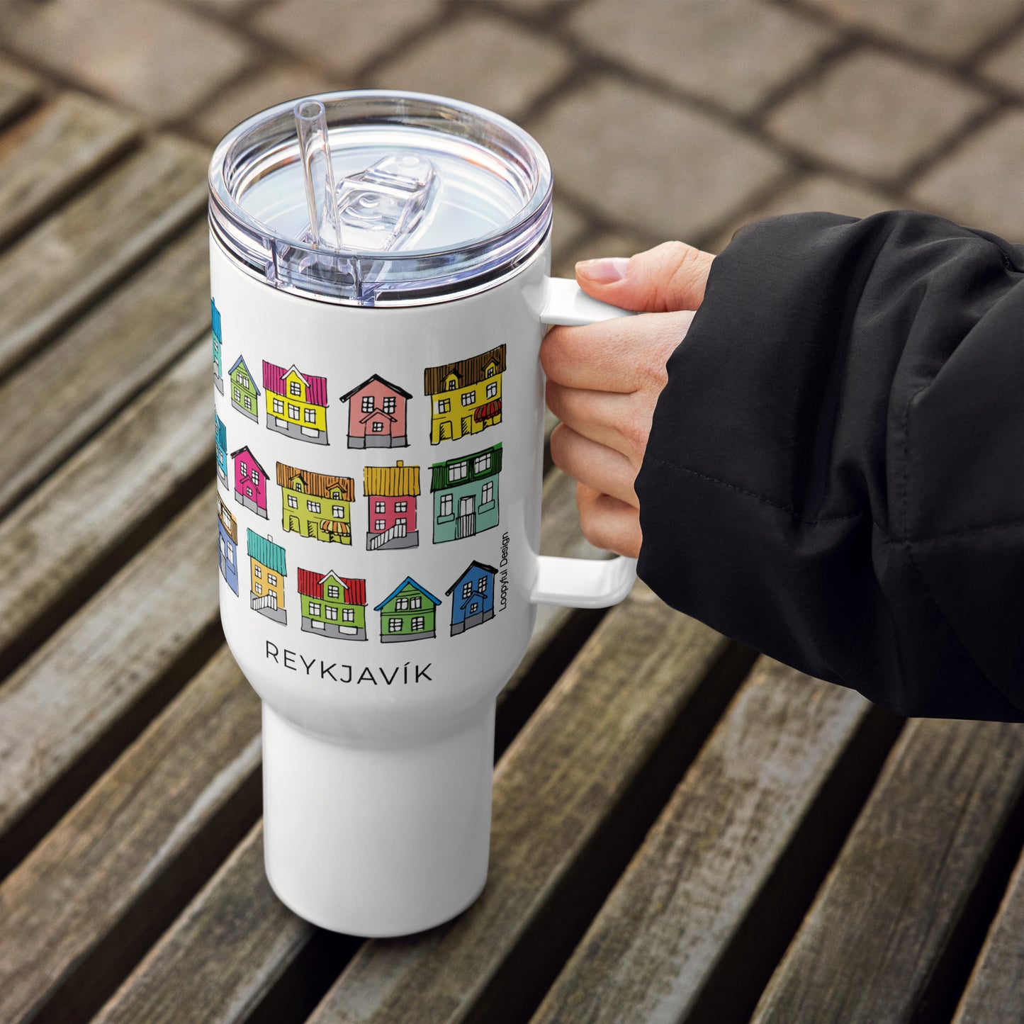 White travel mug with colorful house design and 'Reykjavök' text, held by a person on a wooden surface.